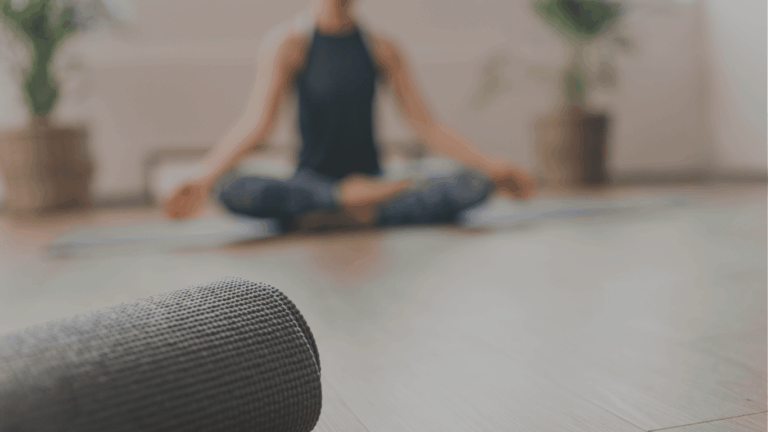 Person meditiert im Schneidersitz, im Vordergrund eine gerollte Yogamatte